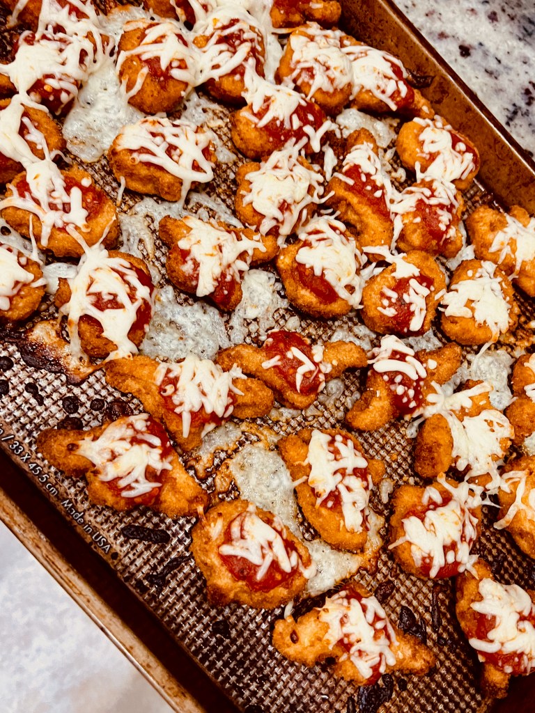 A baking sheet, covered with chicken nuggets sprinkled with tomato sauce and cheese. 